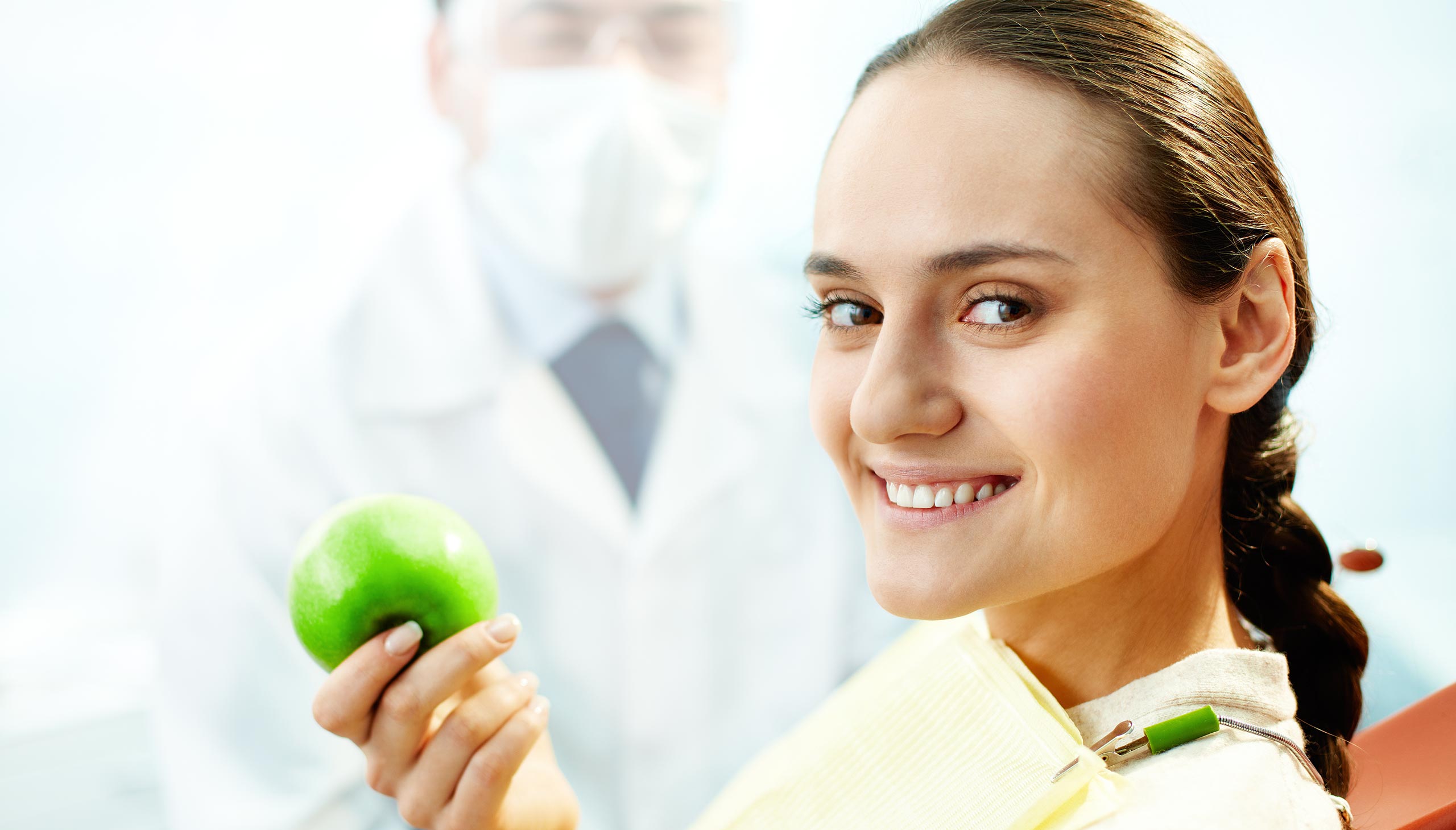 person holds an apple and smiling with a dentist in the background